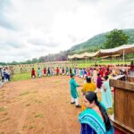 Outdoor cultural event with people in colorful clothes forming a circle while soldiers stand in formation on the left tents and an audience under canopies to the right